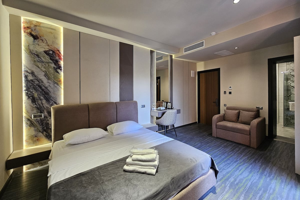 Quarto de hotel moderno com uma cama grande, área de estar, escrivaninha e banheiro com chuveiro.