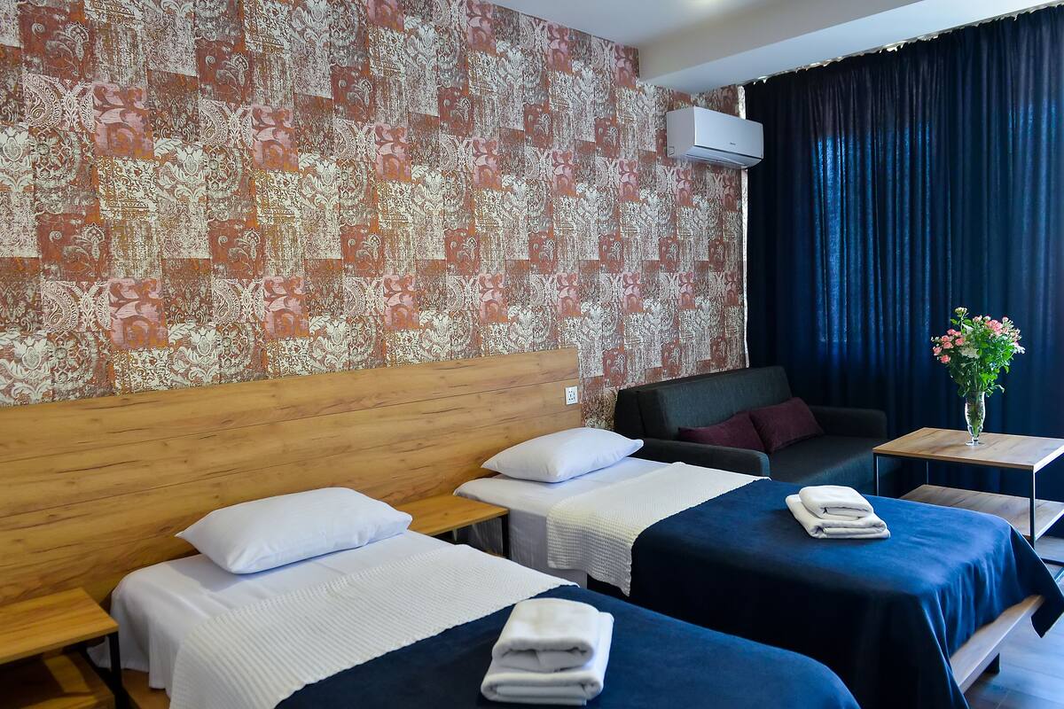 Quarto de hotel com duas camas, um sofá, uma mesa pequena e um detalhe decorativo na parede.