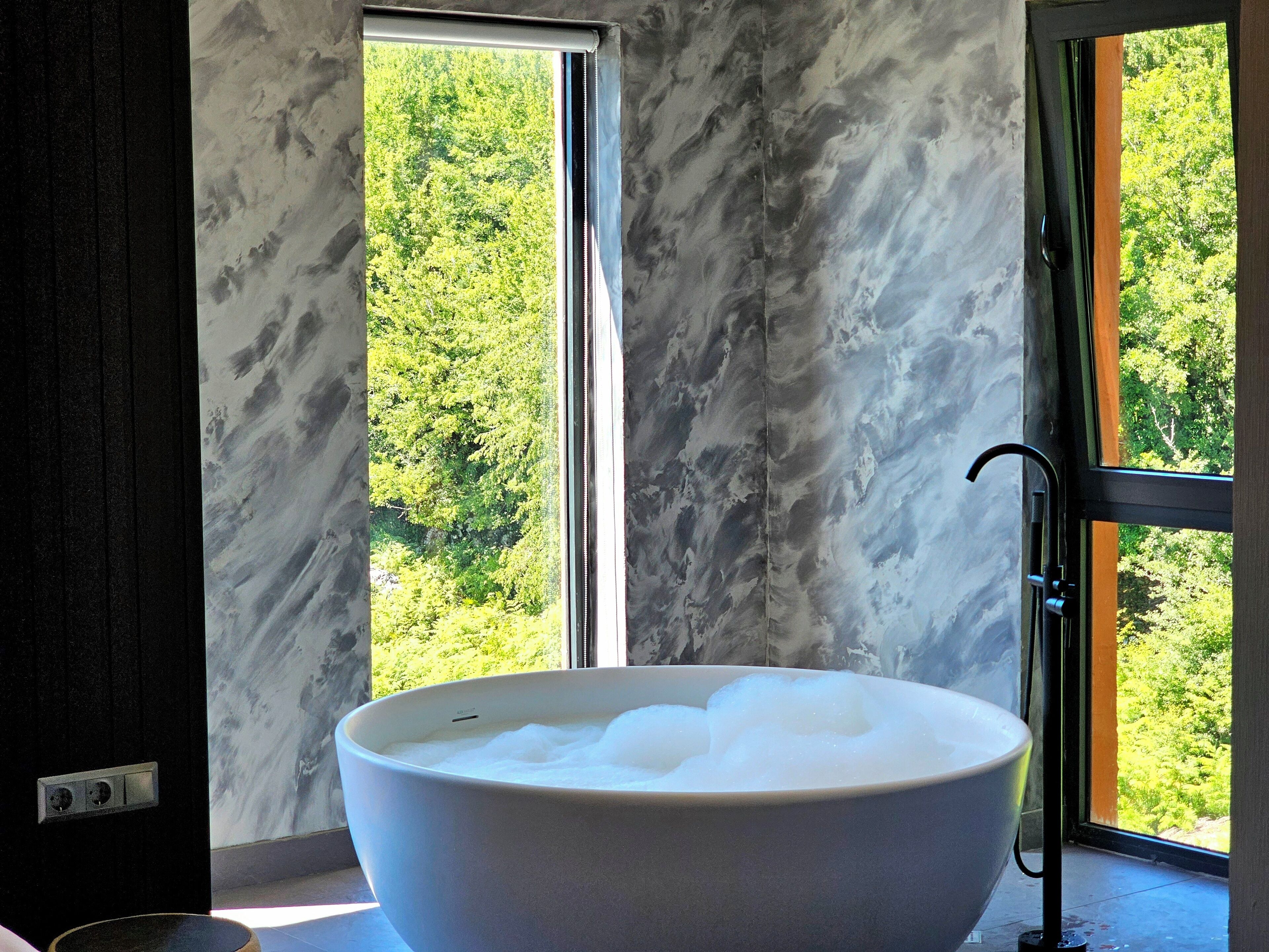 Una bañera blanca grande llena de burbujas, ubicada junto a una ventana con vistas a un bosque.