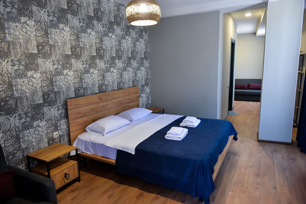 Cama bem arrumada com roupa de cama branca e azul, uma mesinha de cabeceira de madeira e um papel de parede estampado.
