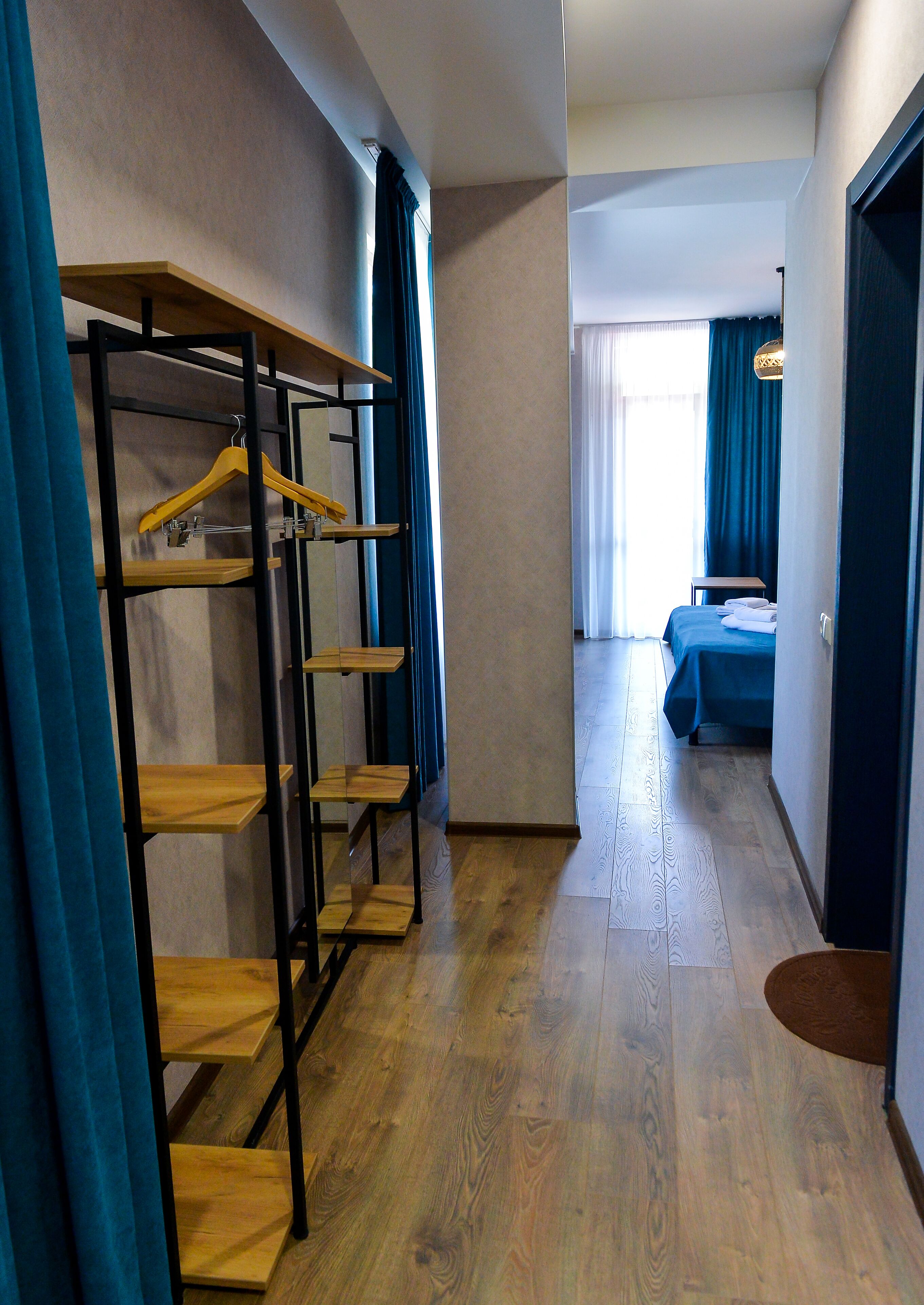 Habitació d'hotel amb sòl de fusta, un llit amb llençol blau, un armari amb prestatges de fusta i un passadís amb cortines blaves.