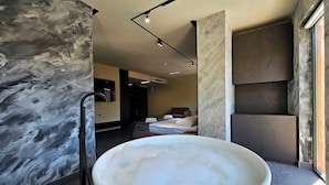 Un baño moderno con una bañera grande, paredes de mármol y vistas a un dormitorio con cama y sillón.