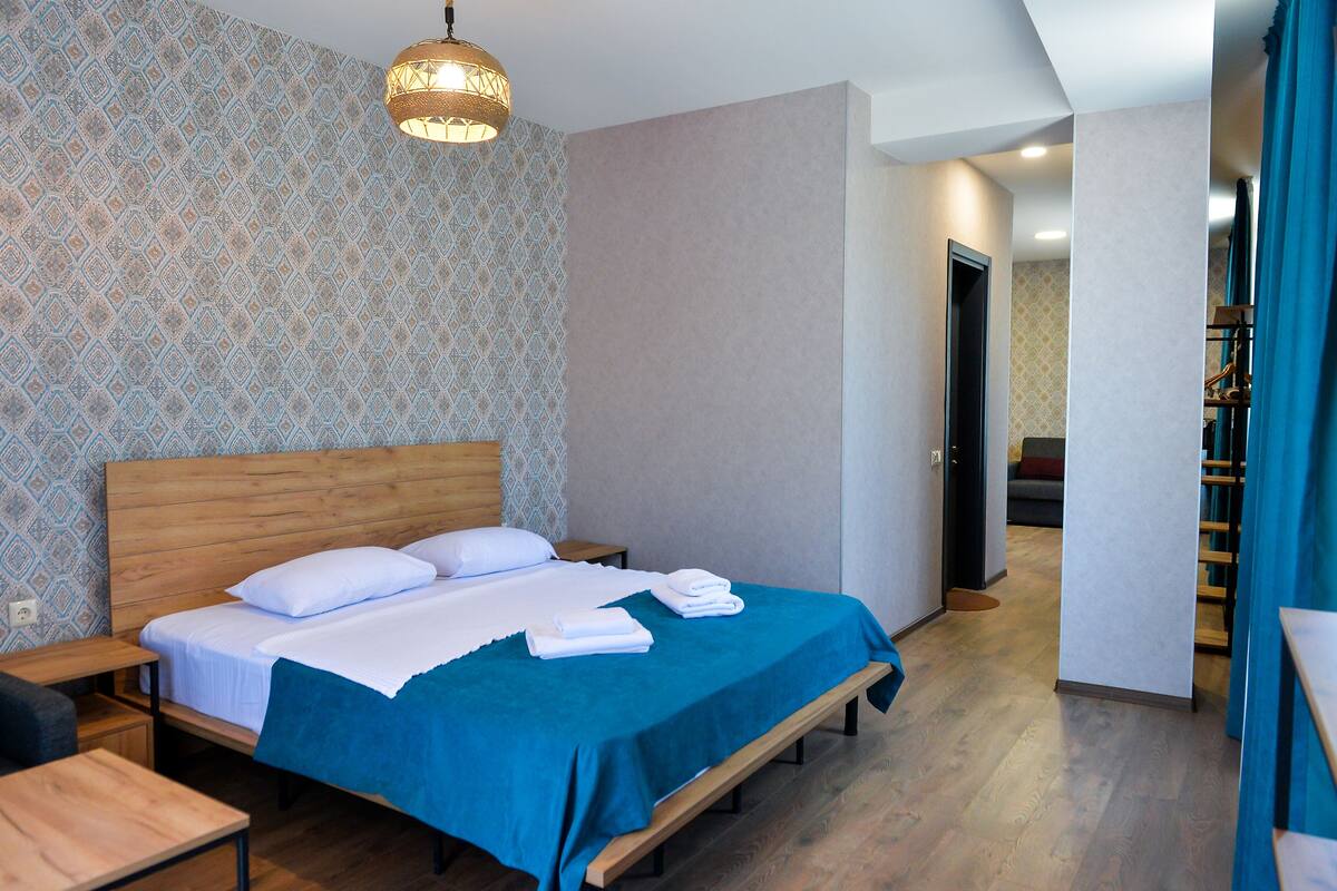 Quarto de hotel com cama de madeira, colcha azul, parede estampada e luminária pendente.