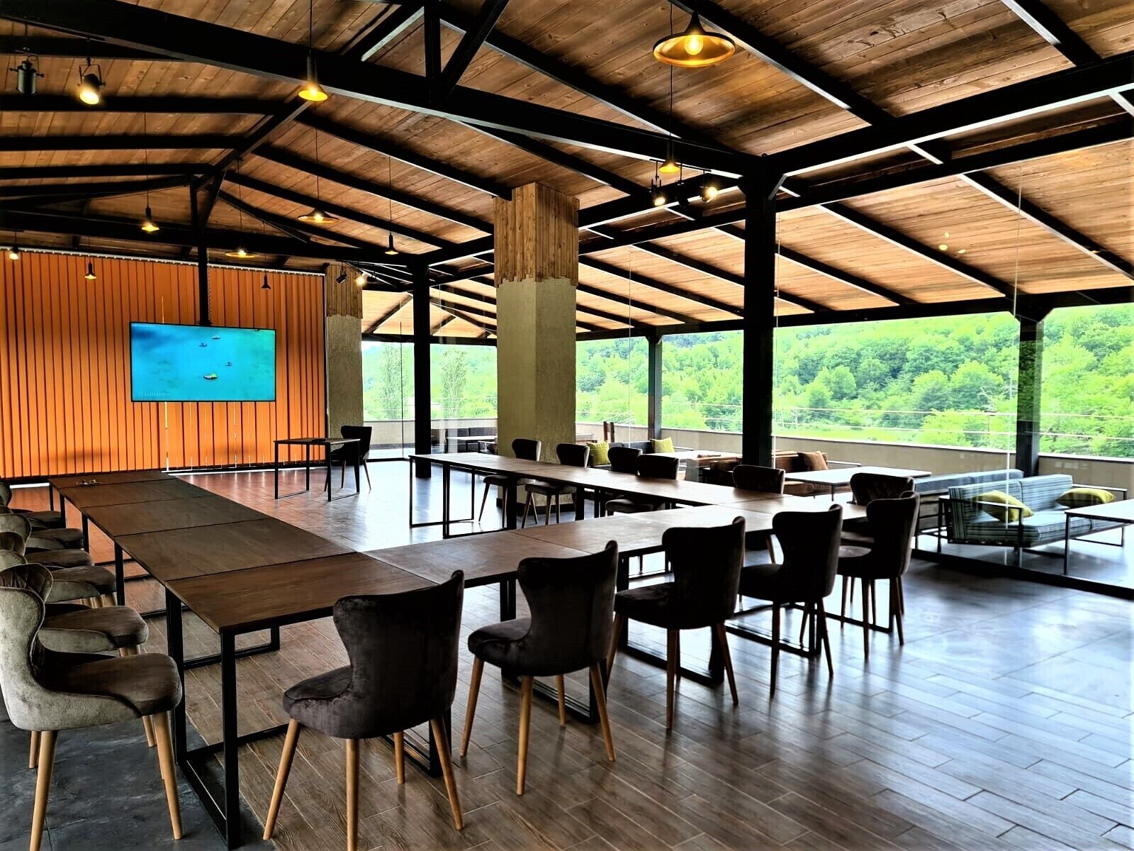 A spacious indoor area with wooden tables and chairs, a large screen on the wall, and large windows offering a view of greenery.
