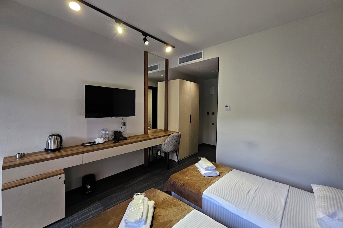 Quarto de hotel com cama, escrivaninha, televisão de tela plana, espelho, guarda-roupa e banheiro visível por uma porta aberta.