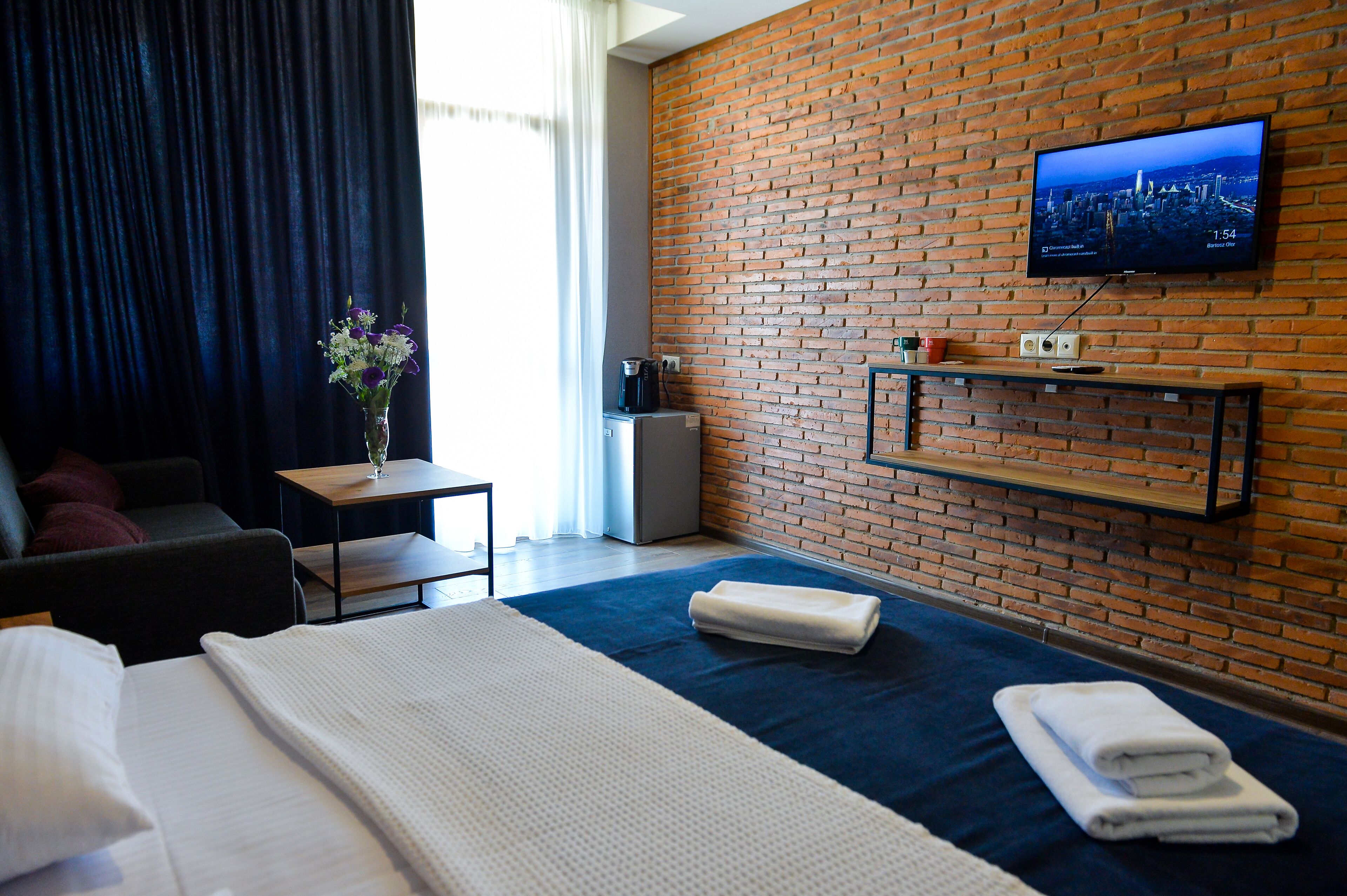 A hotel room with a brick wall, a flat-screen TV, a bed with white linens, a small table with a vase of flowers, and a coffee machine.