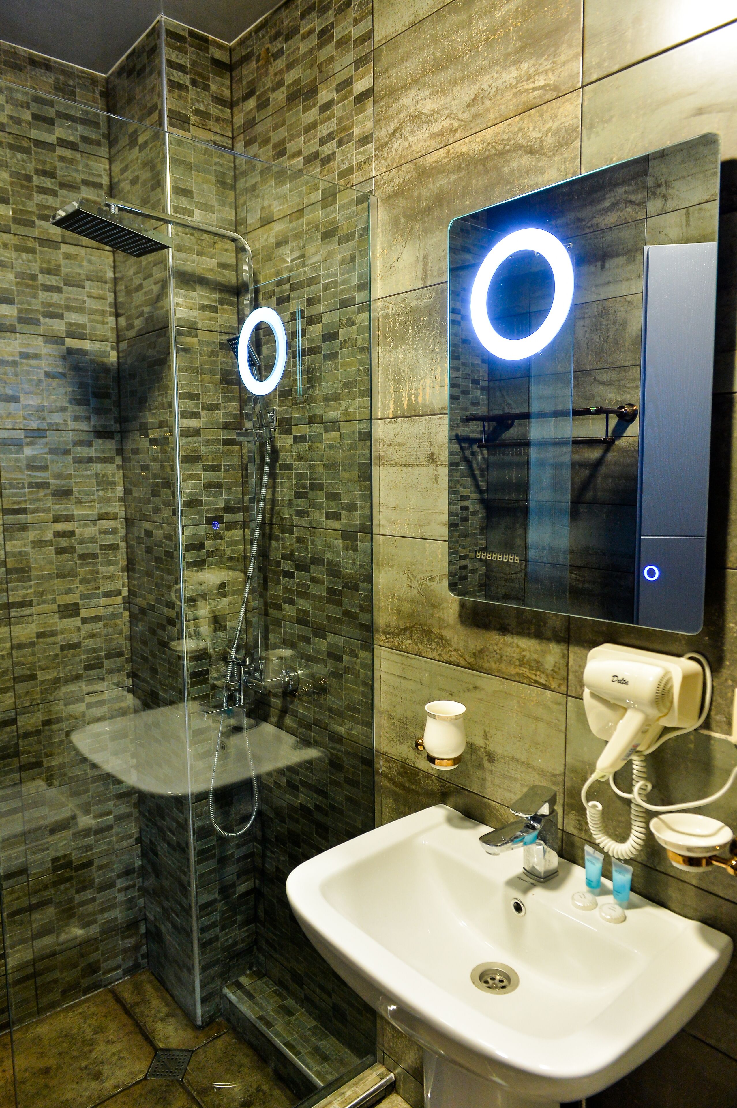 Un baño moderno con ducha de vidrio, lavamanos blanco y espejo con luz circular.