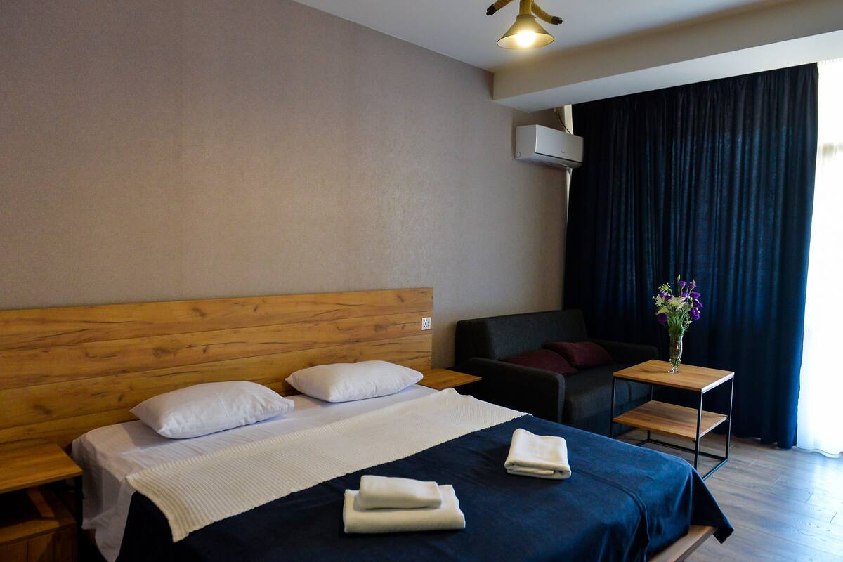 Quarto de hotel com cama de madeira, uma pequena mesa lateral, cortina azul escura e um vaso com flores.