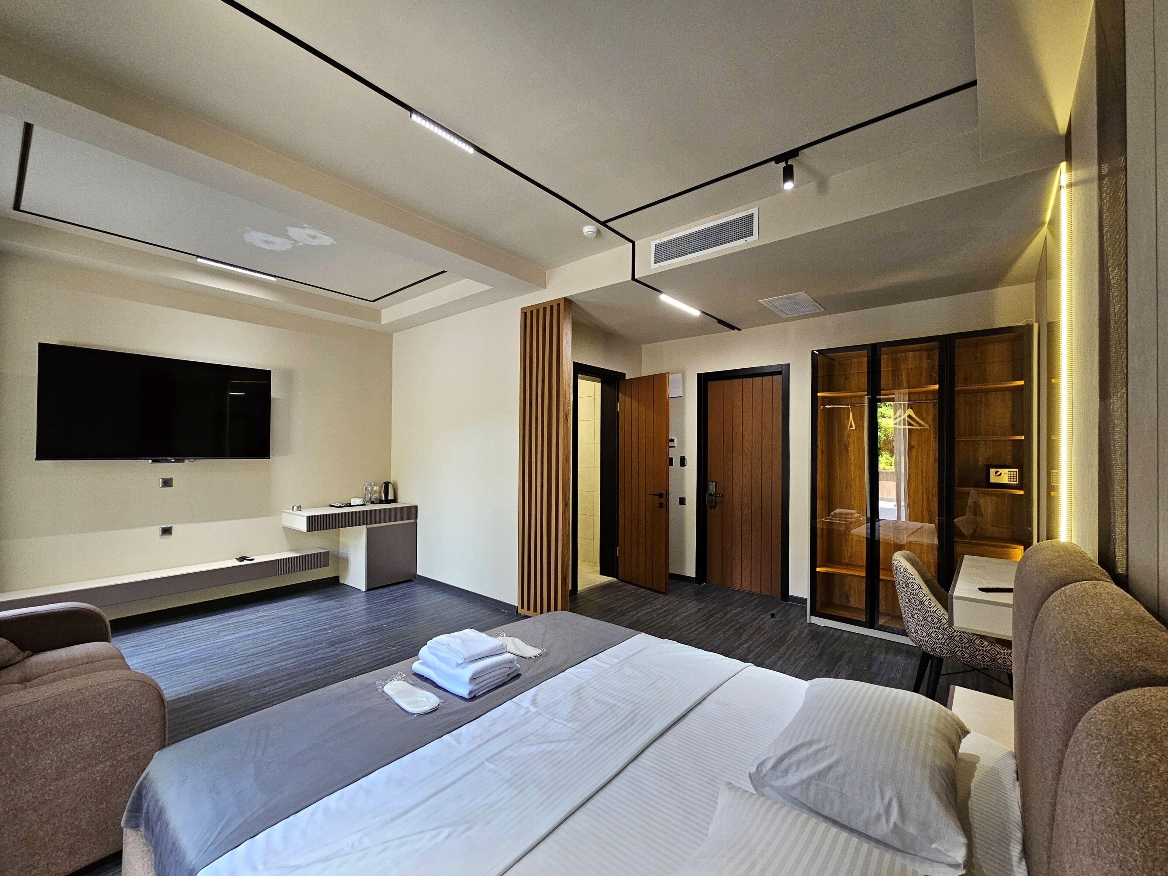 Ein modernes Hotelzimmer mit einem großen Bett, einem flachbildfernseher an der Wand, einem kleinen Schreibtisch mit Stuhl, einem Kleiderschrank und einem begehbaren Kleiderschrank.