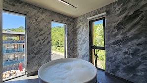 Un baño moderno con una bañera grande llena de burbujas, paredes de mármol y ventanas amplias que ofrecen vistas a un balcón y vegetación exterior.