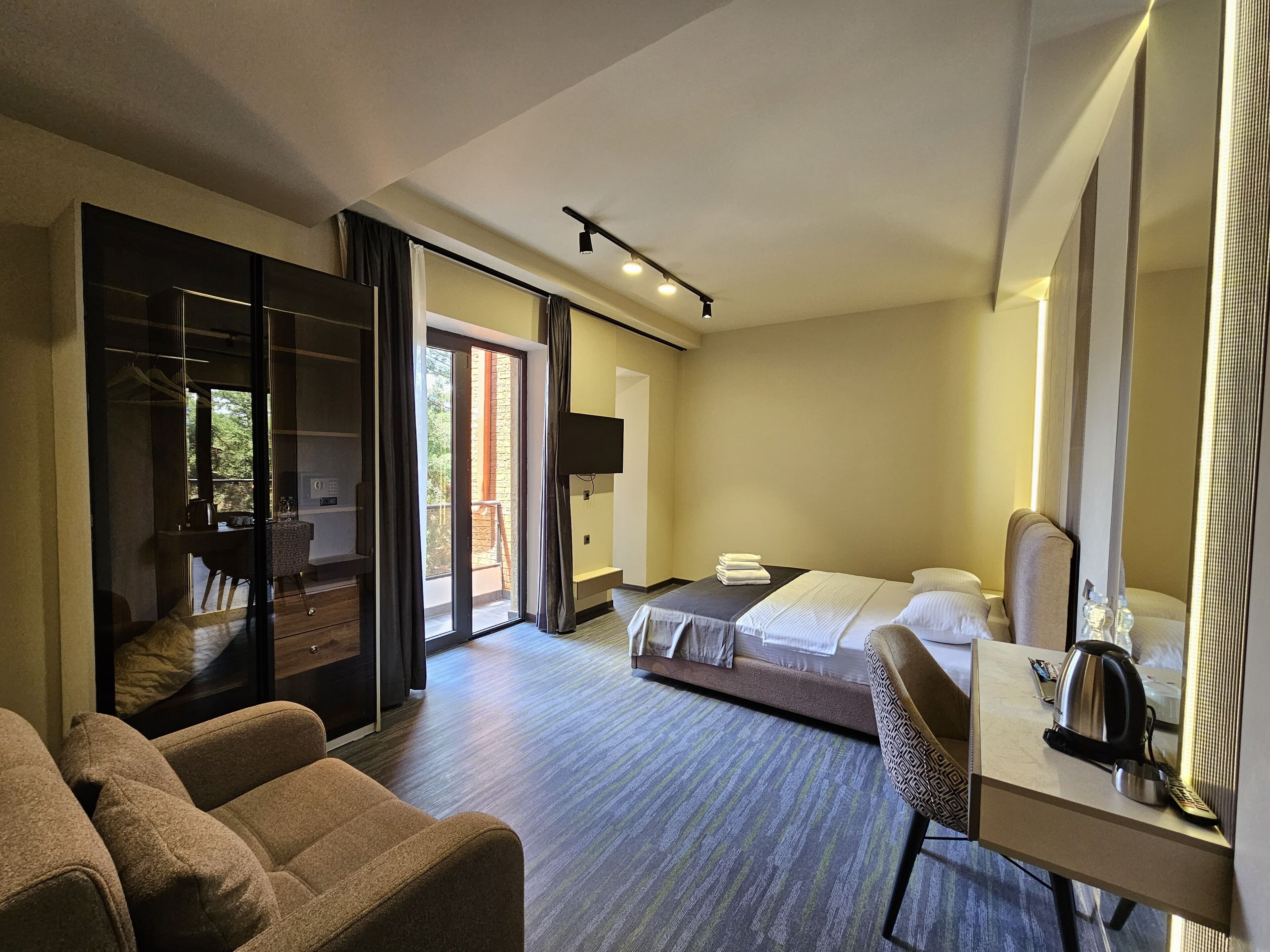 A hotel room with a bed, a desk, a chair, a wardrobe, and a sliding glass door leading to a balcony.