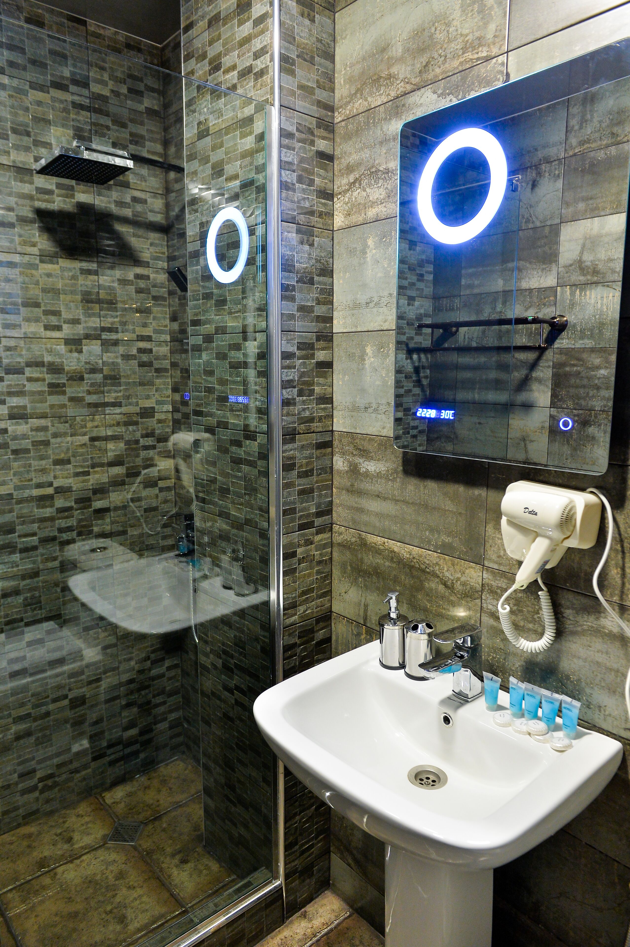 Een moderne badkamer met een douchekabinet van glas, een witte wastafel met toiletartikelen en een digitale spiegel waarop de temperatuur staat.