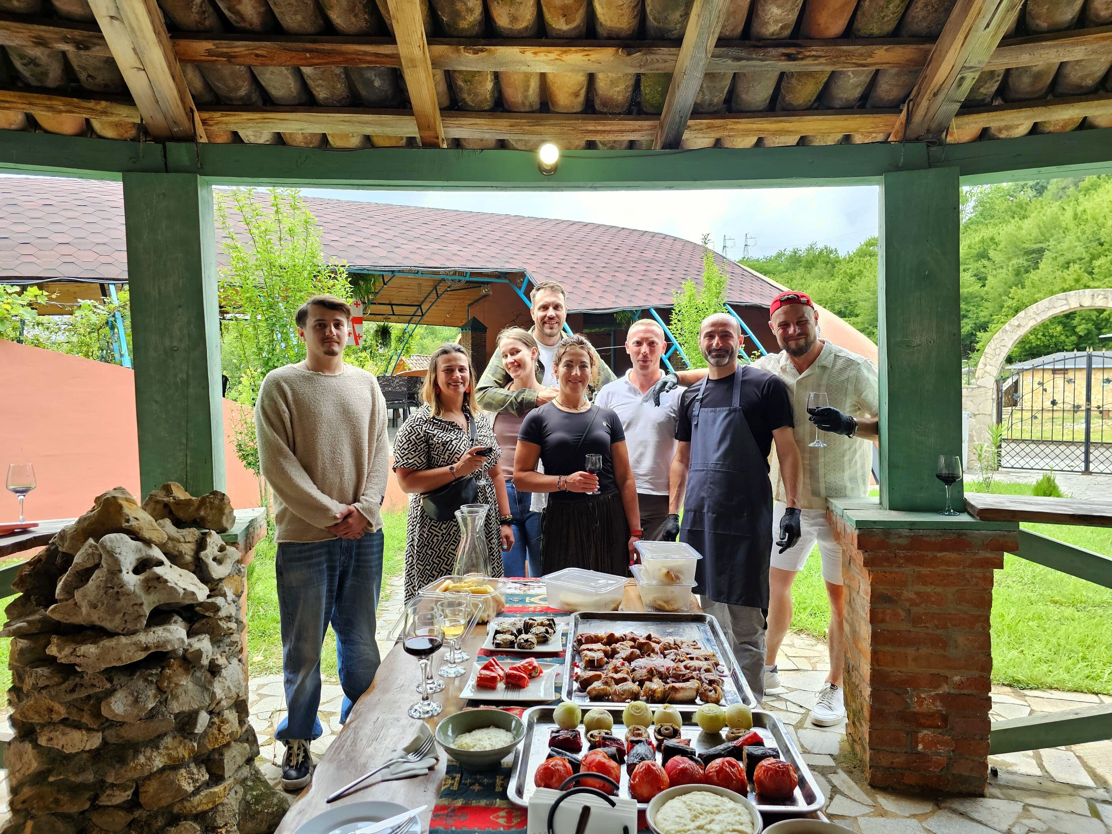 Un grup de persones reunides al voltant d'una taula amb menjar i begudes, sota una estructura de fusta amb un marc verd.