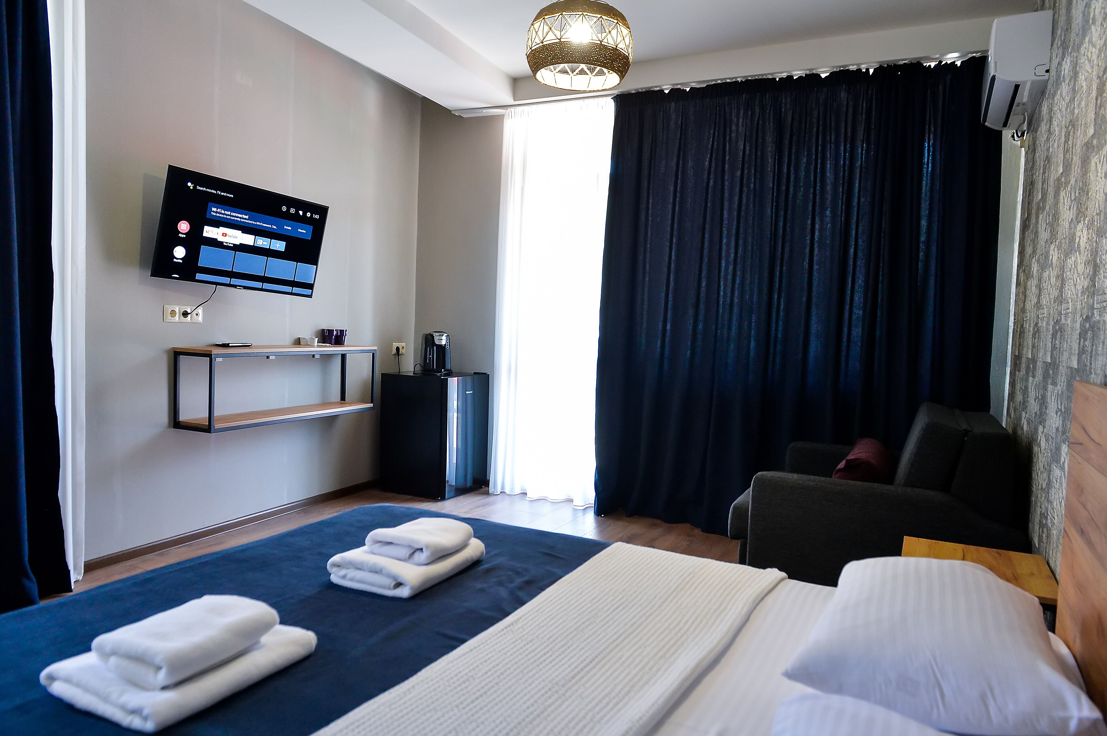 A hotel room with a bed, a TV mounted on the wall, a desk, a chair, and a large window with curtains.