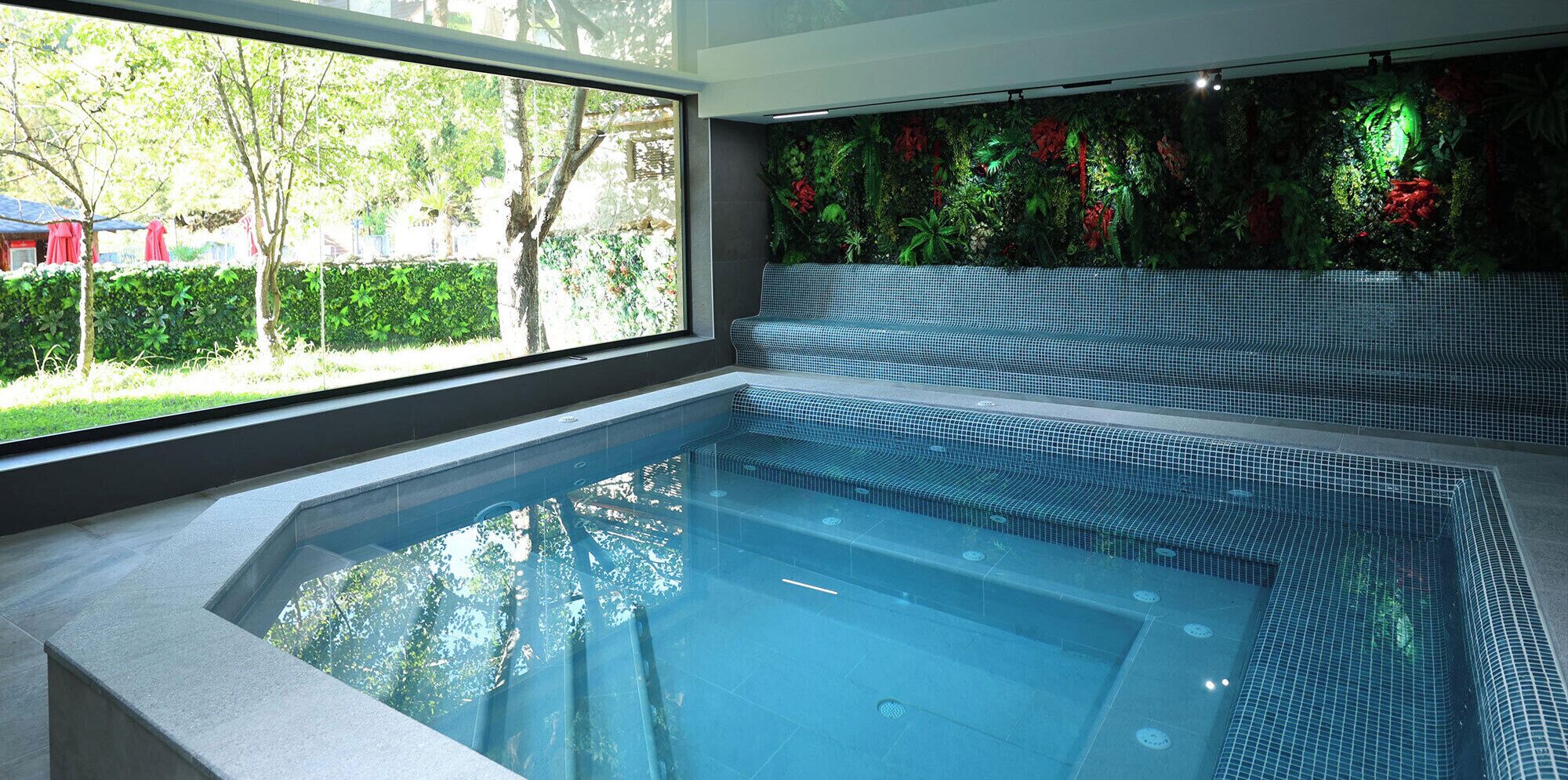 Une piscine intérieure moderne avec des parois en verre transparent, un sol carrelé et une vue sur la verdure environnante.