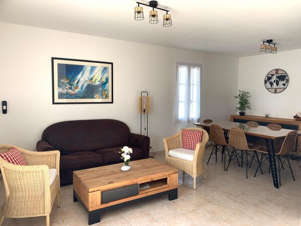 A living room with a brown sofa, wicker chairs, a wooden coffee table, and a dining area with a table and chairs.