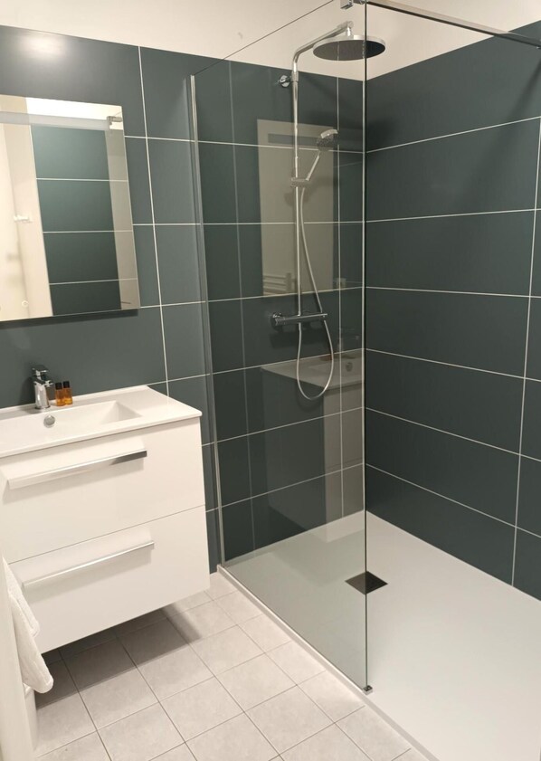 Un baño moderno con ducha envuelta en vidrio, tocador blanco con espejo y paredes de azulejos oscuros.