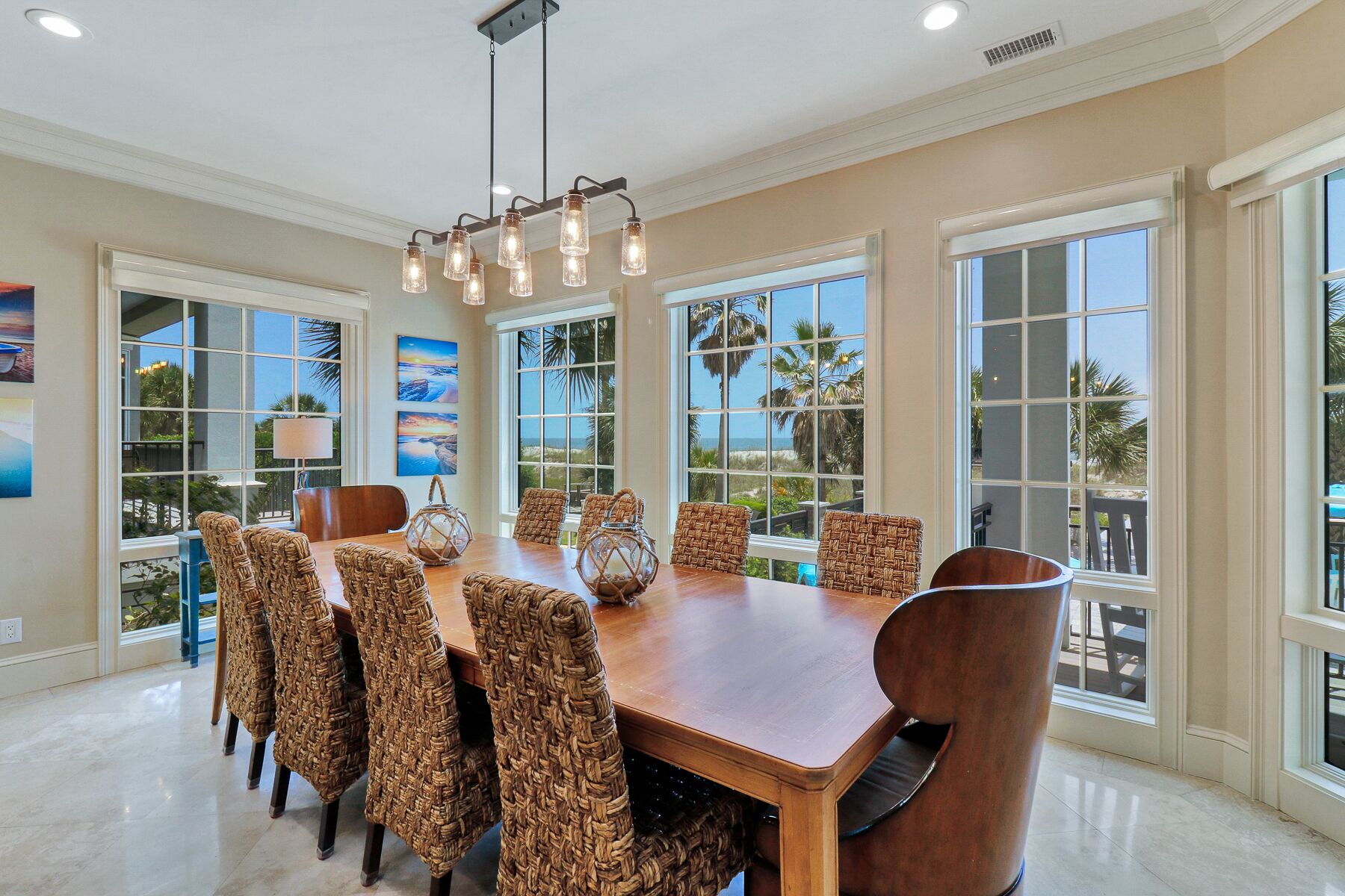 Comedor con mesa de madera, sillas tejidas, ventanas amplias con vistas a palmeras y una lámpara colgante.