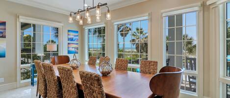 Comedor con mesa de madera, sillas tejidas, ventanas amplias con vistas a palmeras y una lámpara colgante.