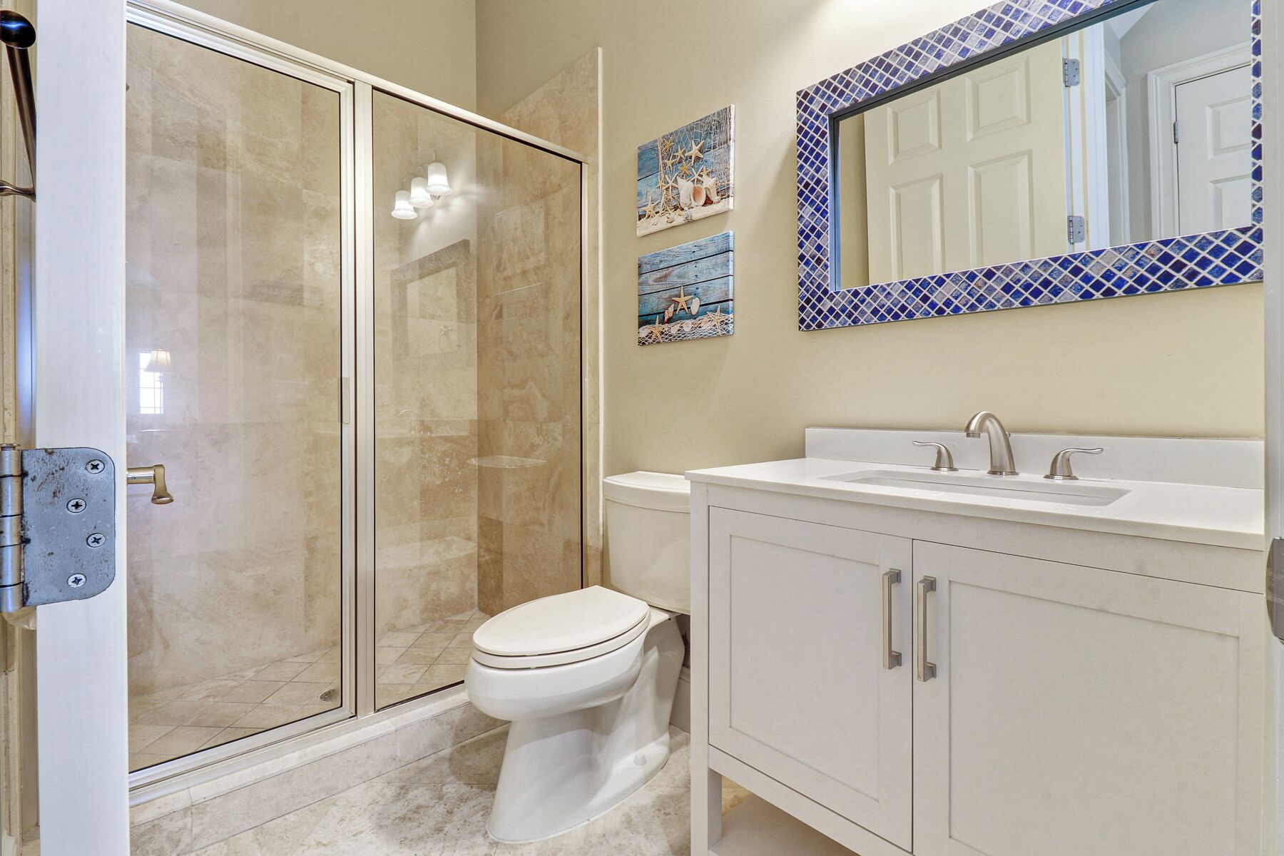 A bathroom with a glass shower enclosure, a toilet, a sink with a mirror above it, and two wall-mounted decorative items.