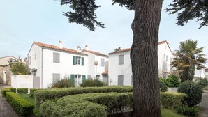A white building with green shutters, surrounded by well-maintained hedges and a large tree. - House TELLINE      . (Saint-Martin-de-Ré)
