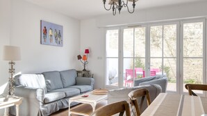 A living room with a grey sofa, a dining table with chairs, a chandelier, and a large window with a view of the outside. - House TELLINE      . (Saint-Martin-de-Ré)