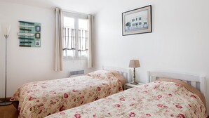 A room with two beds, a window with curtains, a framed picture on the wall, and a signboard with destinations. - House TELLINE      . (Saint-Martin-de-Ré)