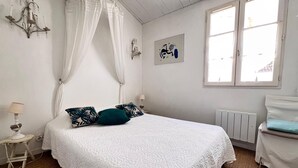 Een slaapkamer met een groot bed, witte gordijnen, een radiator en een raam met gordijnen.