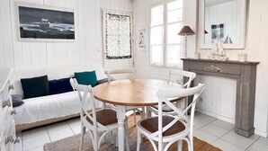 Een eetgedeelte met een ronde houten tafel, witte stoelen, een witte bank met blauwgroene kussens en een open haard met een scheepsdecoratie.