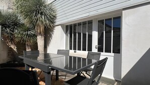 A patio with a glass table and chairs, a palm tree, and a white building with large windows. - House ANNEXE       . (Saint-Martin-de-Ré)