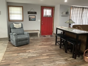 A living room with a grey armchair, a red door, a dining table with two chairs, and a lamp. - Country Cottage (West Union)