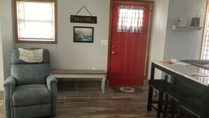 A room with a red door, a welcome sign, a framed picture, a grey armchair, and a dining table with chairs.