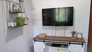 A room with a mounted TV, a white cabinet with a wooden top, a plant, and a small shelf with decorative items.