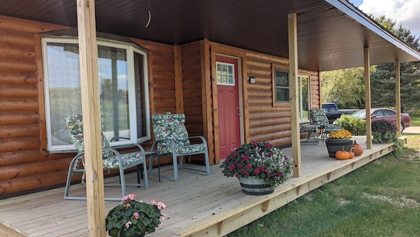 A log cabin with a porch, two chairs, a red door, and potted plants.