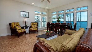 A spacious living room with a wooden floor, a ceiling fan, and large windows offering a view of the outdoors. - 🌞😎Pelican Roost - 4 Bedroom Bayfront Home in Fort Morgan!😎🌞 (Gulf Shores)