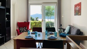 Un coin repas avec une table dressée pour quatre personnes, une chaise rouge, une chaise noire et un canapé gris. Un lustre est suspendu au plafond, et une fenêtre offre une vue sur la verdure et les montagnes.