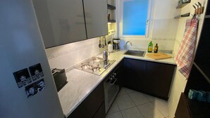 Une cuisine de taille réduite, équipée d’une cuisinière à gaz, d’un évier et d’une fenêtre avec des stores.