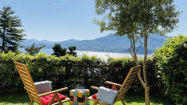 Deux chaises en bois avec des coussins, une petite table et un coussin coloré, le tout installé sur une pelouse verdoyante avec vue sur des montagnes et un lac.