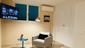 Un salon avec un téléviseur fixé au mur, un fauteuil, une petite table avec une plante et une lampe.