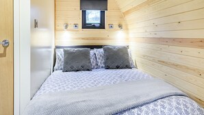 A bedroom with a wooden sloped ceiling, a bed with patterned bedding, two bedside lamps, and a window above the bed. - A beautiful and very comfortable glamping experience. (East Haddon)