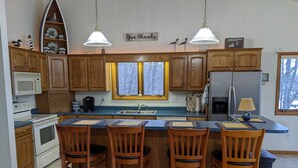 Una cocina con armarios de madera, una isla central con taburetes de bar, microondas, refrigerador y una ventana con vistas a árboles.