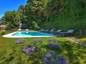 Piscina con sillas de descanso y flores púrpuras en un jardín ajardinado.