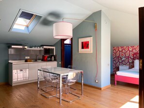 Una habitación compacta con kitchenette, una mesa de comedor con sillas, una cama con cabecera estampada y un tragaluz.