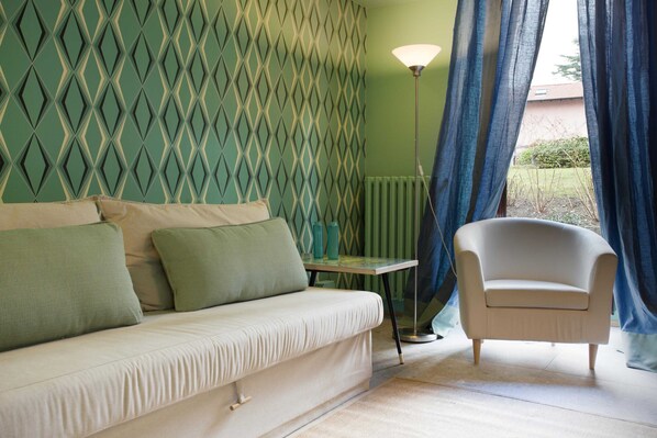 Una habitación con pared de diseño verde, un sofá blanco con cojines verdes, una mesita, una lámpara y una silla.