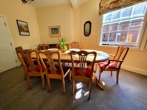A dining room with a round wooden table surrounded by six chairs with red cushions. There is a vase with flowers on the table, and a framed picture on the wall.
