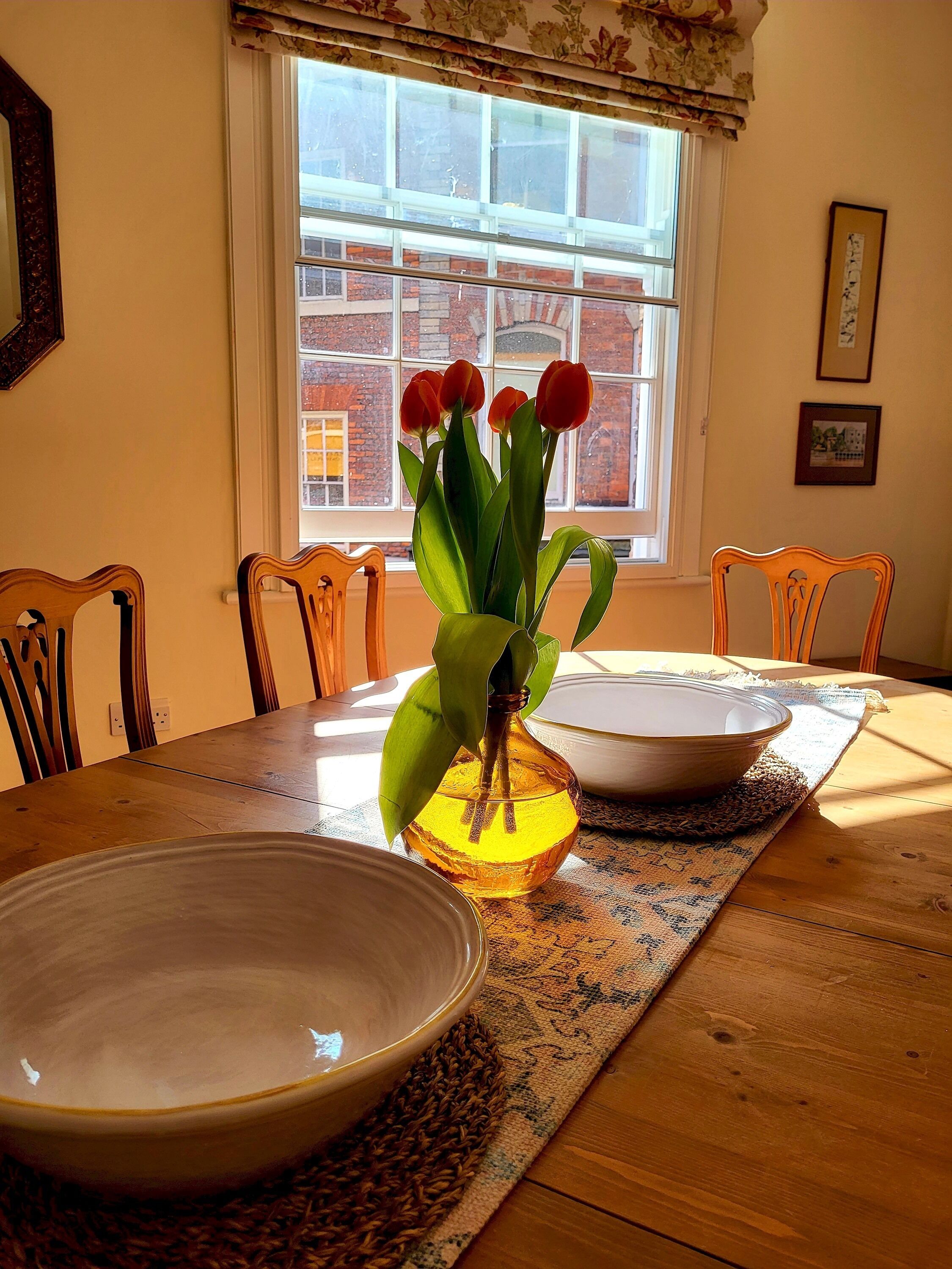 A dining table with a bowl, a vase with flowers, and a window with a view of a brick building.