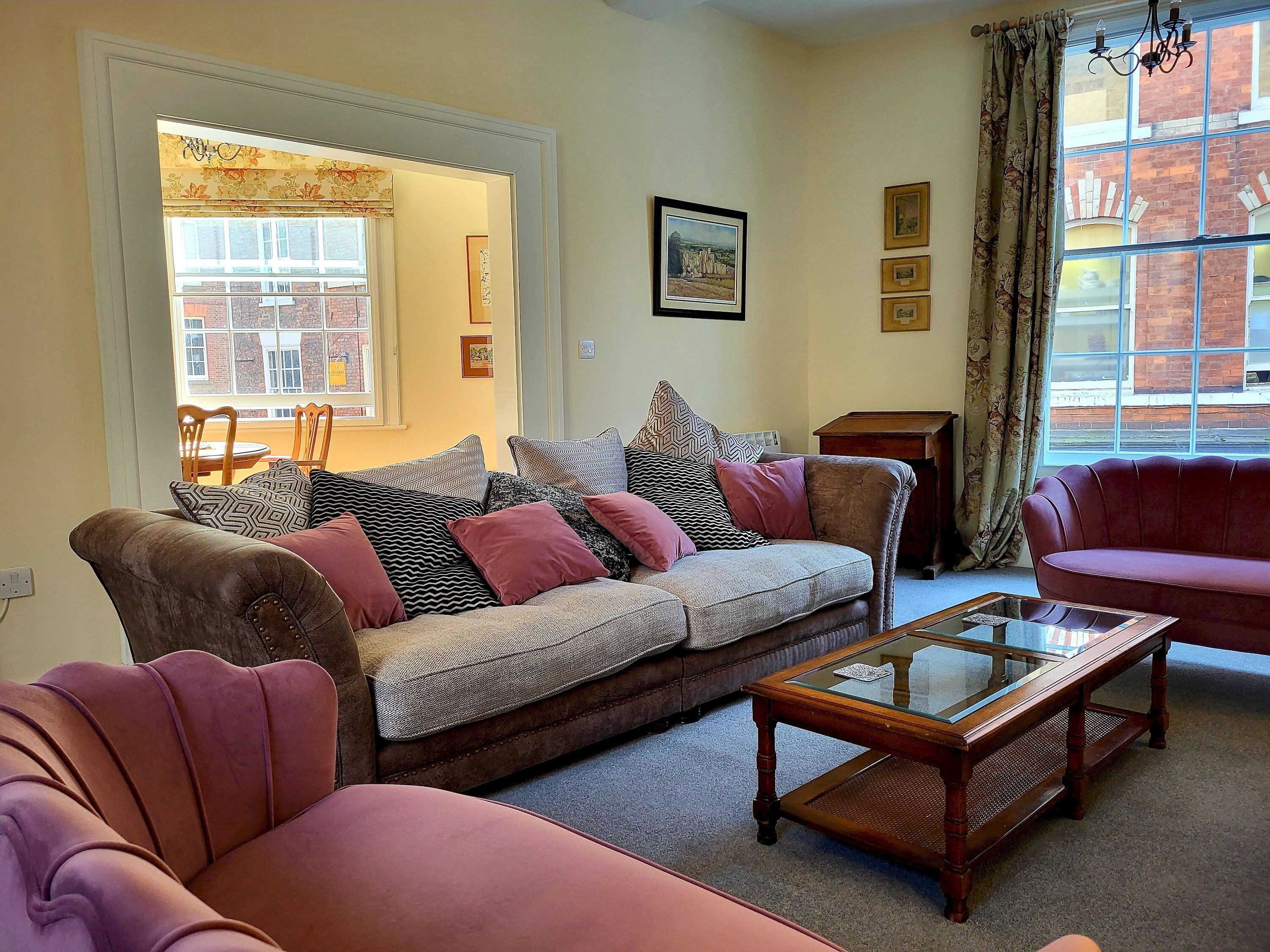 A living room with a sofa, armchairs, a glass-top coffee table, and a view of a brick building through a window.