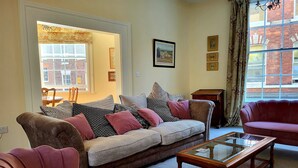 Ein Wohnzimmer mit einer Couch, Sesseln, einem Couchtisch mit Glasplatte und Blick auf ein Ziegelgebäude durch ein Fenster.