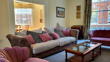 A living room with a sofa, armchairs, a glass-top coffee table, and a view of a brick building through a window.