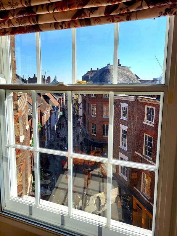 Blick durch ein Fenster mit floralem Vorhang. Man sieht eine schmale Gasse mit Backsteingebäuden, ein Geschäft und Fußgänger.
